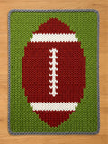 A knitted chunky blanket featuring a football design. The football is red with white stripes and stitching, set against a green background. The blanket has a gray border and is made of a textured yarn.