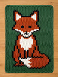 May include: A knitted chunky blanket featuring a fox design. The fox is primarily orange, with white and black accents, set against a green background. The blanket has a thick, textured appearance.