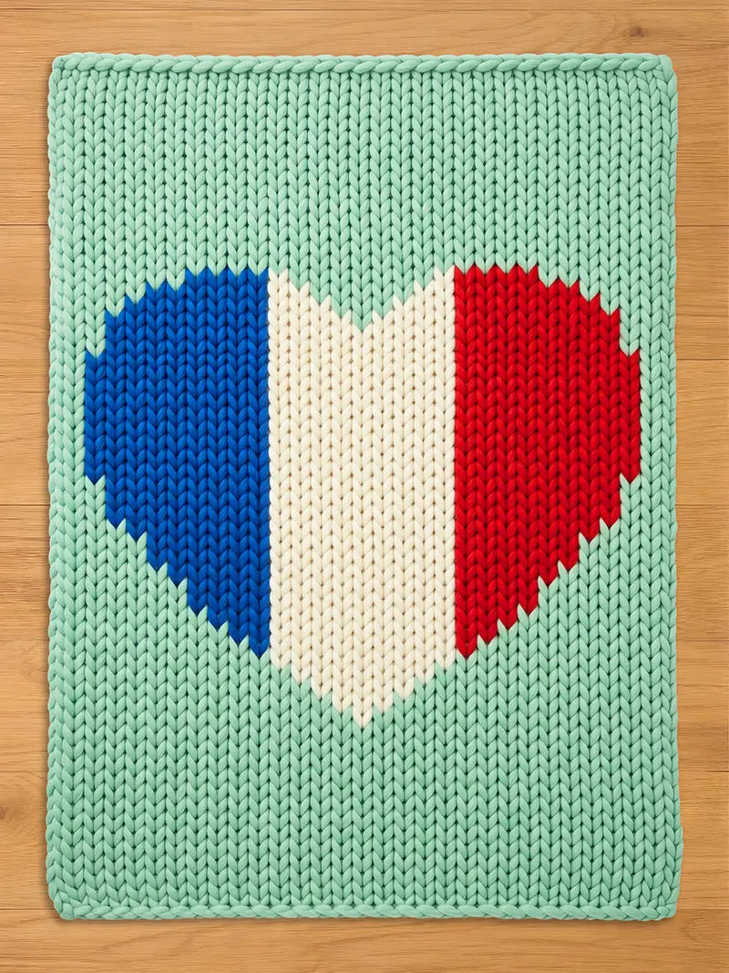 A knitted rectangular chunky blanket with a heart-shaped design in the colors of the French flag: blue, white, and red. The background is a light green color. The blanket is made of thick yarn.