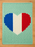 A knitted rectangular chunky blanket with a heart-shaped design in the colors of the French flag: blue, white, and red. The background is a light green color. The blanket is made of thick yarn.