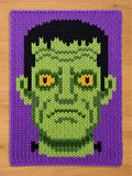 A chunky knit blanket featuring a green Frankenstein monster face with yellow eyes, black hair, and a purple background.