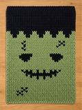 A rectangular knitted blanket featuring a green face with black hair, black eyes, a jagged black stitched mouth, and black stitched scars on the forehead and lower cheek. The knit pattern is thick and uniform throughout the piece.