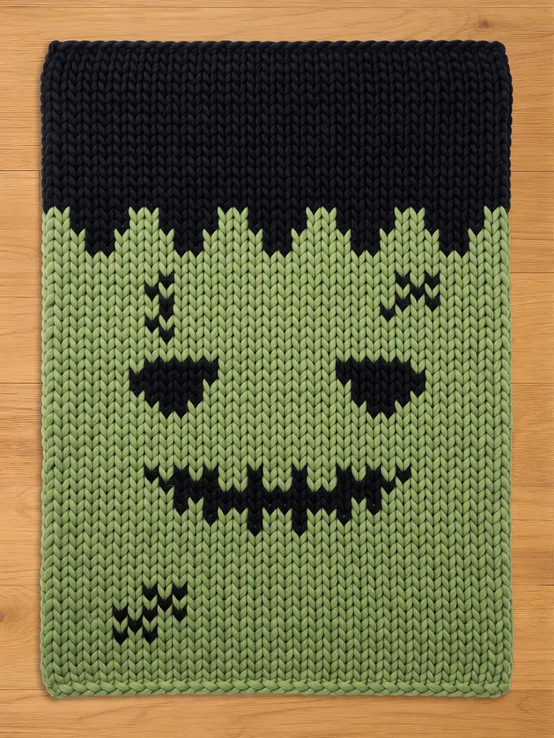 A rectangular knitted blanket featuring a green face with black hair, black eyes, a jagged black stitched mouth, and black stitched scars on the forehead and lower cheek. The knit pattern is thick and uniform throughout the piece.