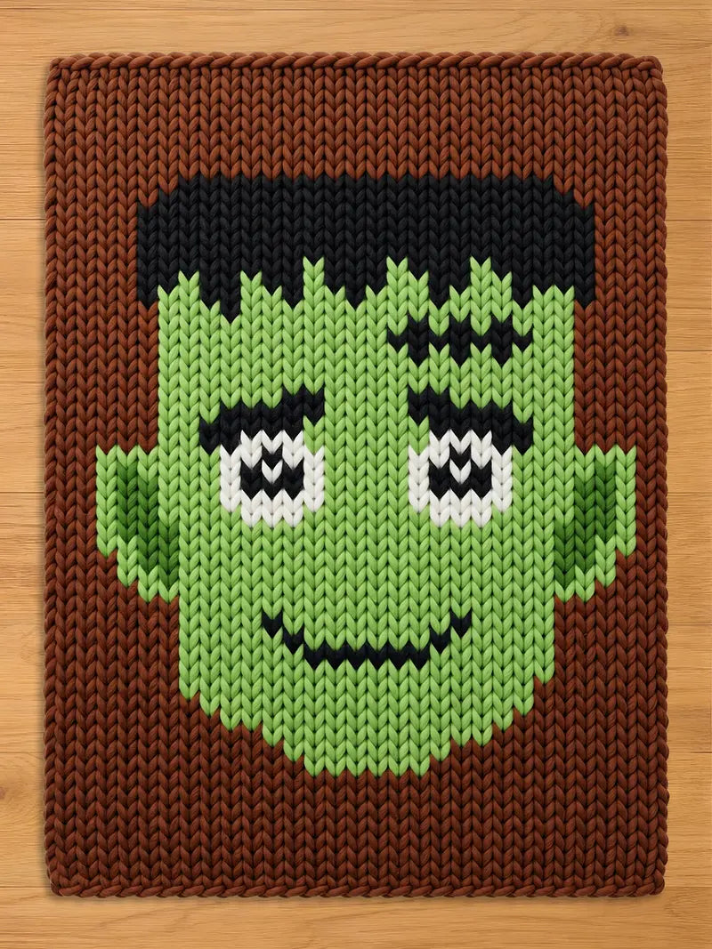A rectangular knitted blanket featuring a graphic of a green-skinned character with black hair, thick black eyebrows, white eyes with black pupils, and a black stitched mouth, all set against a solid brown background.