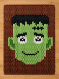 A rectangular knitted blanket featuring a graphic of a green-skinned character with black hair, thick black eyebrows, white eyes with black pupils, and a black stitched mouth, all set against a solid brown background.