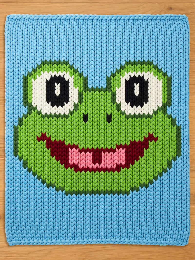 A knitted chunky blanket featuring a smiling frog face. The frog's face is green with white and black eyes, a pink mouth, and a dark red tongue. The background is light blue, and the edges are neatly finished.