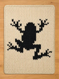 Off-white knitted chunky blanket featuring a large black frog design. The frog is centrally placed, with outstretched limbs. The blanket has a thick, textured appearance, suitable for a nursery or children's room.