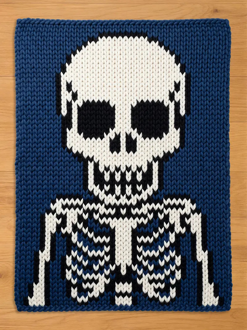 a chunky rectangular knit blanket featuring a graphic of a human skeleton. The design is created with white, black, and dark blue yarn, showcasing a skull with hollow eye sockets and a rib cage structure against a solid dark blue background.