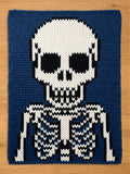 a chunky rectangular knit blanket featuring a graphic of a human skeleton. The design is created with white, black, and dark blue yarn, showcasing a skull with hollow eye sockets and a rib cage structure against a solid dark blue background.