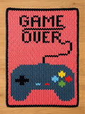A knitted chunky knit blanket with a pink background and a black border. The blanket features a blue video game controller at the bottom and the words "GAME OVER" in black at the top. The controller has colorful buttons.