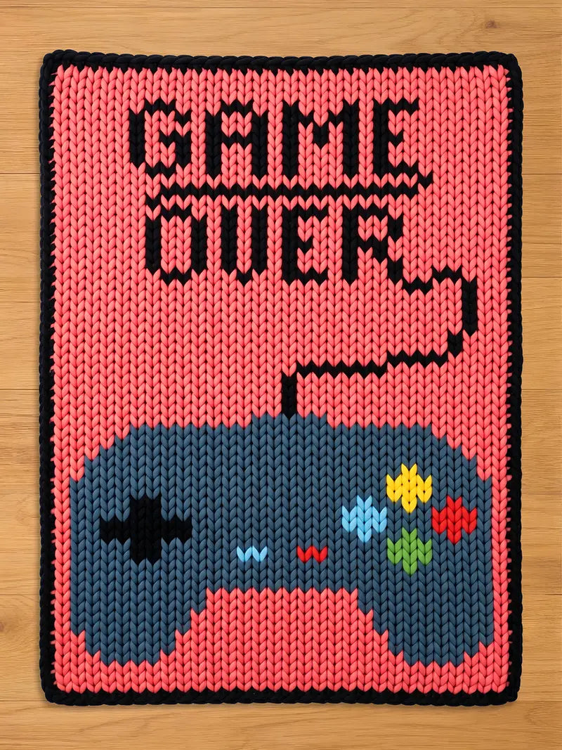 A knitted chunky knit blanket with a pink background and a black border. The blanket features a blue video game controller at the bottom and the words "GAME OVER" in black at the top. The controller has colorful buttons.