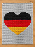A knitted blanket featuring a heart design in the colors of the German flag: black, red, and yellow. The heart is centered on a gray knitted background. The blanket is made of soft, textured material.