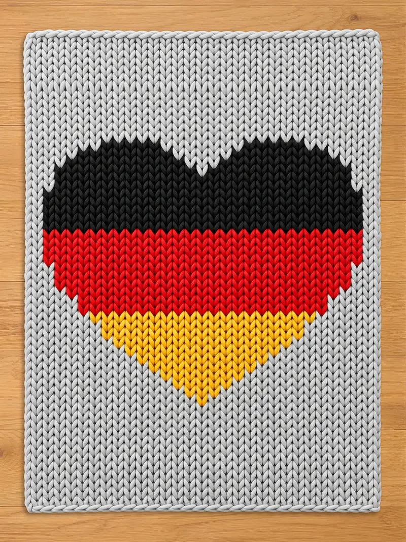 A knitted blanket featuring a heart design in the colors of the German flag: black, red, and yellow. The heart is centered on a gray knitted background. The blanket is made of soft, textured material.