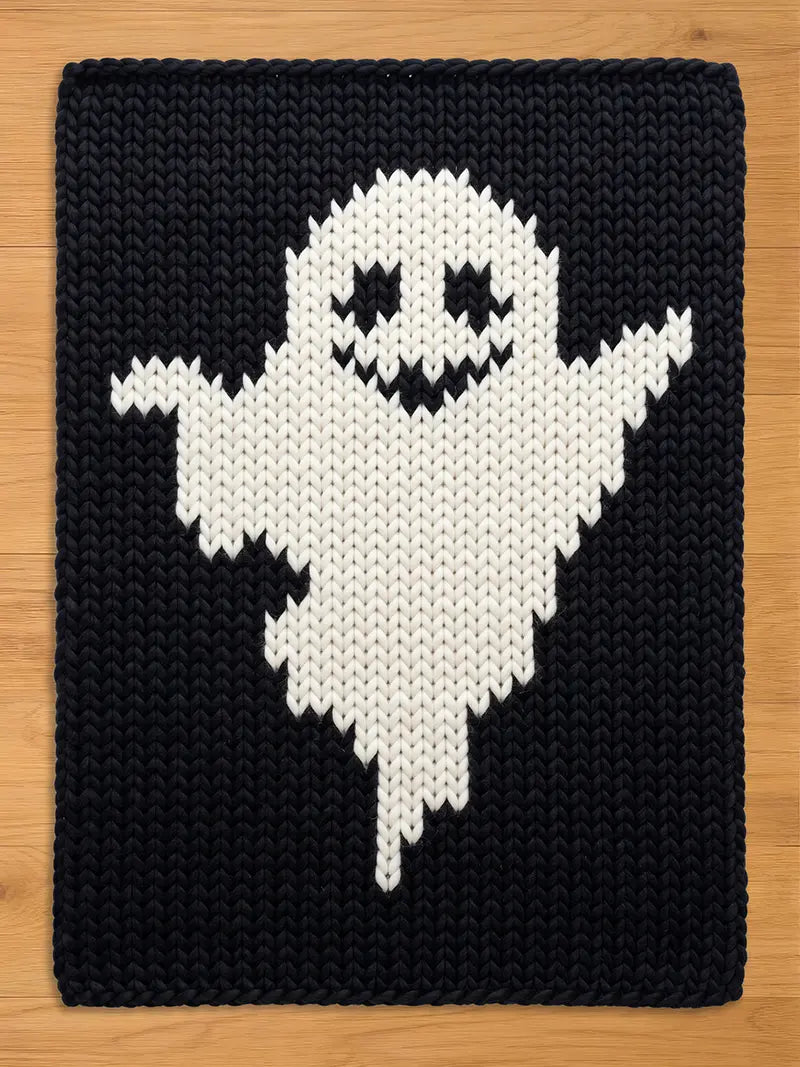 A rectangular knitted blanket featuring a large, white ghost figure centered against a solid black background. The ghost has two triangular eyes and a jagged, wide mouth, all rendered in black yarn within the white knitted shape.