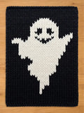 A rectangular knitted blanket featuring a large, white ghost figure centered against a solid black background. The ghost has two triangular eyes and a jagged, wide mouth, all rendered in black yarn within the white knitted shape.
