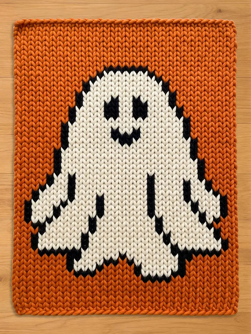 A rectangular knitted blanket featuring a large white ghost with black eyes and a black mouth centered on a solid orange background. The knit pattern is chunky and uniform, with black yarn outlining the ghost shape against the orange.
