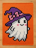 A rectangular knit blanket featuring a vibrant orange background with a central graphic of a white ghost wearing a purple witch hat with pink accents. The ghost has black eyes, a black mouth, and pink cheeks, all rendered in knit.