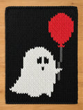 A rectangular knit blanket featuring a black background with a white ghost character in the center. The ghost has two black eyes and a small black mouth, and it is holding a red balloon attached to a grey string.
