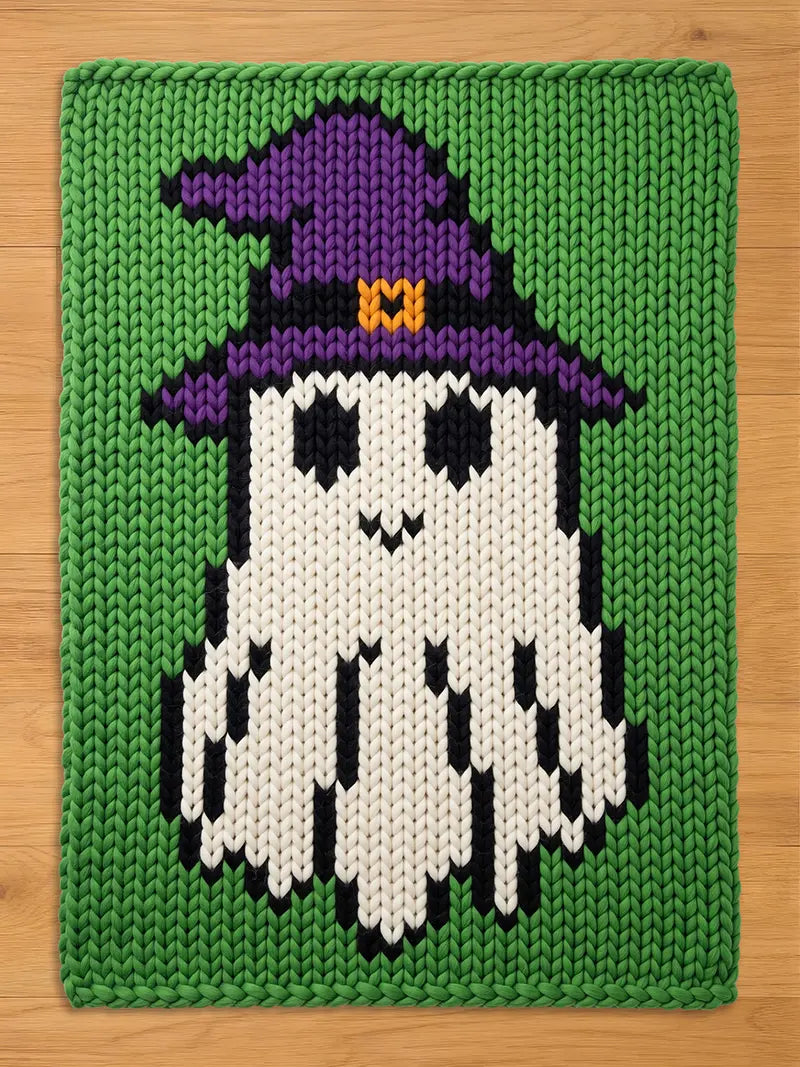A rectangular chunky knit blanket featuring a white ghost character wearing a purple witch hat with an orange buckle. The ghost is set against a solid green background, with black yarn used for the eyes, mouth, and ghost body outlines.