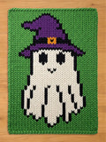 A rectangular chunky knit blanket featuring a white ghost character wearing a purple witch hat with an orange buckle. The ghost is set against a solid green background, with black yarn used for the eyes, mouth, and ghost body outlines.