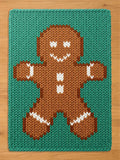 A rectangular knit blanket featuring a brown gingerbread person centered on a teal background. The gingerbread figure has white stitched details for the eyes, mouth, buttons, and cuffs on the arms and legs, created with a chunky yarn texture.