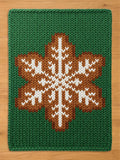 A green chunky knitted rectangular blanket with a large brown and white snowflake design in the center, laid flat on a light wood surface.