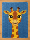 A knitted blue chunky knit blanket featuring a giraffe design. The giraffe has a yellow and brown pattern with black eyes and a smiling mouth. The blanket is made of a thick, textured yarn.