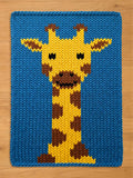 A knitted blue chunky blanket with a yellow and brown giraffe design. The giraffe has brown spots, a yellow face, and black eyes. The background is a solid blue color. 