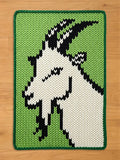 A knitted rectangular chunky blanket featuring a goat's head in black and white against a green background. The blanket has a green border. The goat's head is a pixelated design, giving it a modern, graphic look.