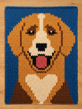 Chunky knitted dog portrait with a blue background. The dog has brown, tan, and white fur, with a pink tongue and black eyes. The dog is smiling. The knitted texture is visible.
