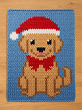 A chunky knit blanket laid flat on a wooden surface, featuring a knitted pattern of a Golden Retriever wearing a red and white Santa hat and a red bow tie against a solid light blue background.