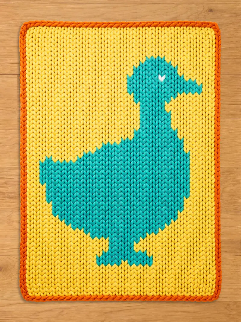 A knitted rectangular chunky blanket featuring a teal duck design on a yellow background. The duck has a white heart detail. The blanket has an orange border and is made of a soft, textured material.
