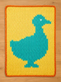 A knitted rectangular chunky blanket featuring a teal duck design on a yellow background. The duck has a white heart detail. The blanket has an orange border and is made of a soft, textured material.