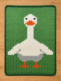 Chunky knit blanket featuring a large white goose pattern on a vibrant green background. The goose has an orange beak, orange webbed feet, and a gray lower body. The blanket showcases a thick, textured yarn knit stitch with a dark green braided border, displayed on a wooden floor.