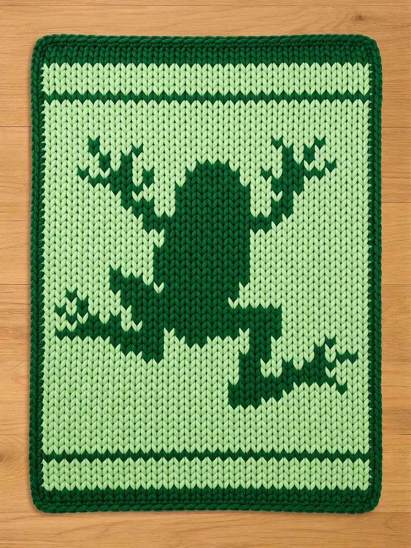 A knitted green frog-themed chunky blanket. The blanket features a dark green frog silhouette on a lighter green background. White and dark green stripes border the top and bottom.