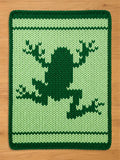 A knitted green frog-themed chunky blanket. The blanket features a dark green frog silhouette on a lighter green background. White and dark green stripes border the top and bottom.