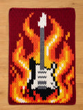 A knitted guitar design with flames in shades of yellow, orange, and red. The guitar is black, white, and gray, set against a burgundy background. The image is a close-up of a knitted item.