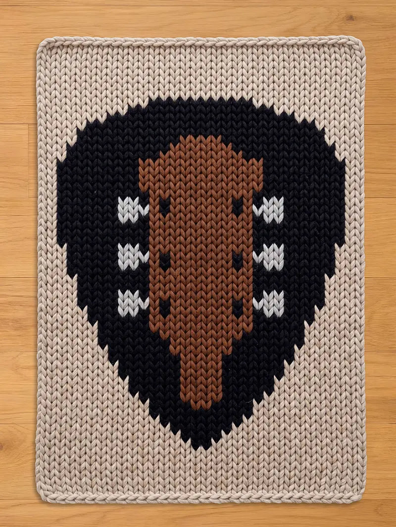 A rectangular, knitted blanket with a guitar design. The guitar is brown, set against a black heart shape, and surrounded by a beige border. The guitar has white accents. The blanket is placed on a wooden surface.