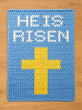 A blue knitted chunky blanket with the words "HE IS RISEN" in white and a yellow cross. The knitted texture is visible, creating a handmade appearance. The design is simple and bold.