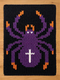 A rectangular knit blanket featuring a large spider design centered on a black background. The spider is primarily purple with orange accents on the leg tips and head, and a white cross-shaped marking on its rounded abdomen.