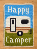 Knitted rectangular chunky blanket with a camper design. The top section is blue with the word "Happy" in white. The camper is white with black details, set on a green field with the word "Camper" in white. The blanket is trimmed with a cream-colored border.