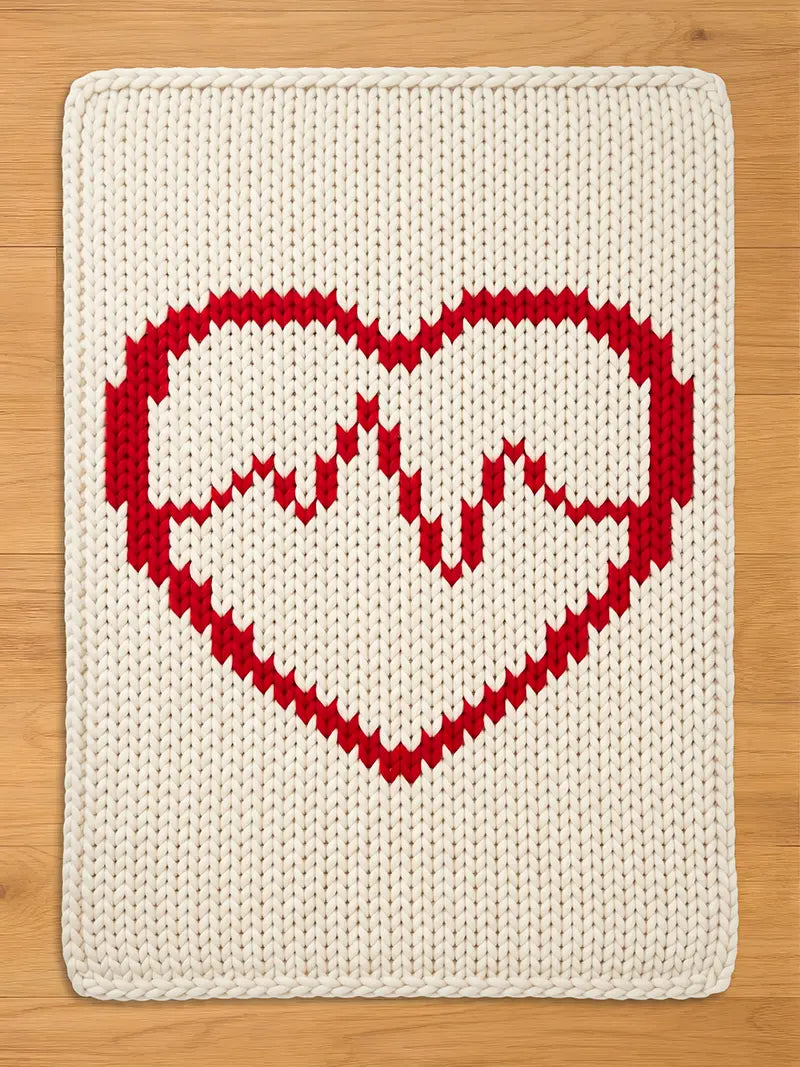 A rectangular cream-colored knit blanket featuring a large red heart design with a red heartbeat line, laid flat on a light wood background.