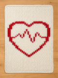 A rectangular cream-colored knit blanket featuring a large red heart design with a red heartbeat line, laid flat on a light wood background.
