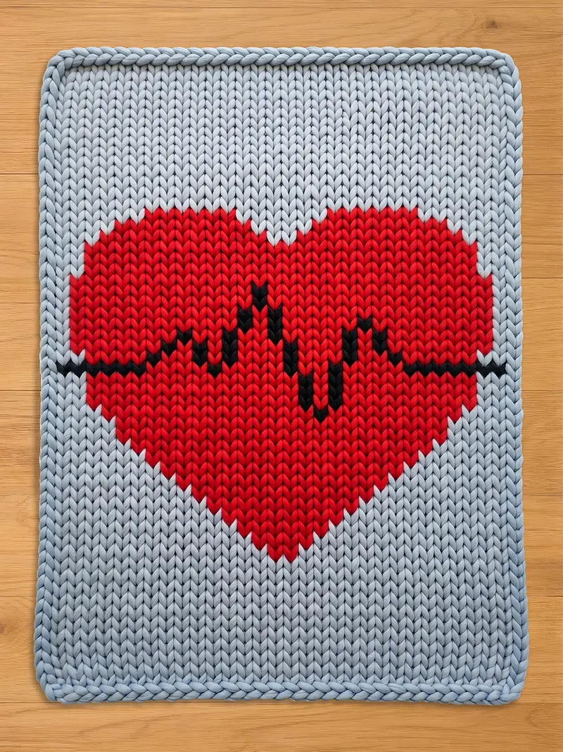 A chunky knit blanket featuring a large red heart with a black heartbeat line across the center on a light gray background, laid flat on a wooden surface.