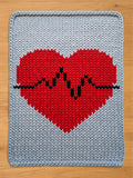 A chunky knit blanket featuring a large red heart with a black heartbeat line across the center on a light gray background, laid flat on a wooden surface.