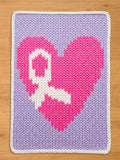 A knitted chunky blanket featuring a pink heart with a white ribbon design on a lavender background. The heart is centered, and the rug has a white border. The blanket is made of yarn.