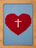 A knitted blanket featuring a large red heart with a white cross in the center, set against a light blue background. The blanket has a gray border and is made of soft, textured yarn.