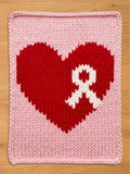 Light pink chunky knit blanket featuring a large red heart with a white awareness ribbon knitted into the center, displayed flat on a wooden surface.