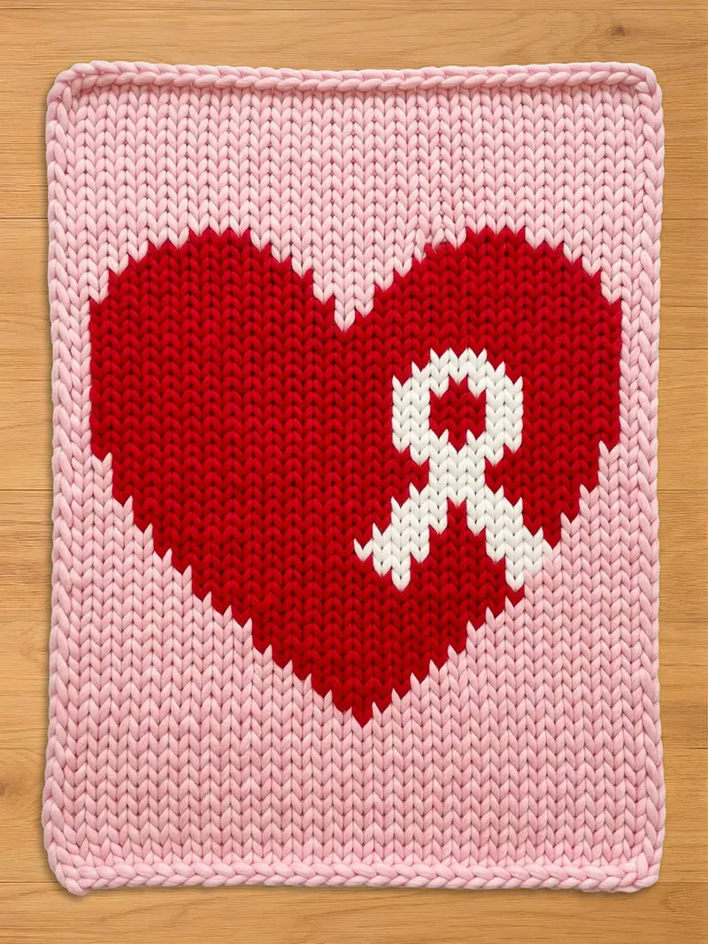 Light pink chunky knit blanket featuring a large red heart with a white awareness ribbon knitted into the center, displayed flat on a wooden surface.