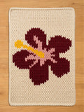 Chunky knit cream blanket featuring a large knit maroon hibiscus flower design with a light pink center and a yellow stamen, displayed flat on a light wood floor.